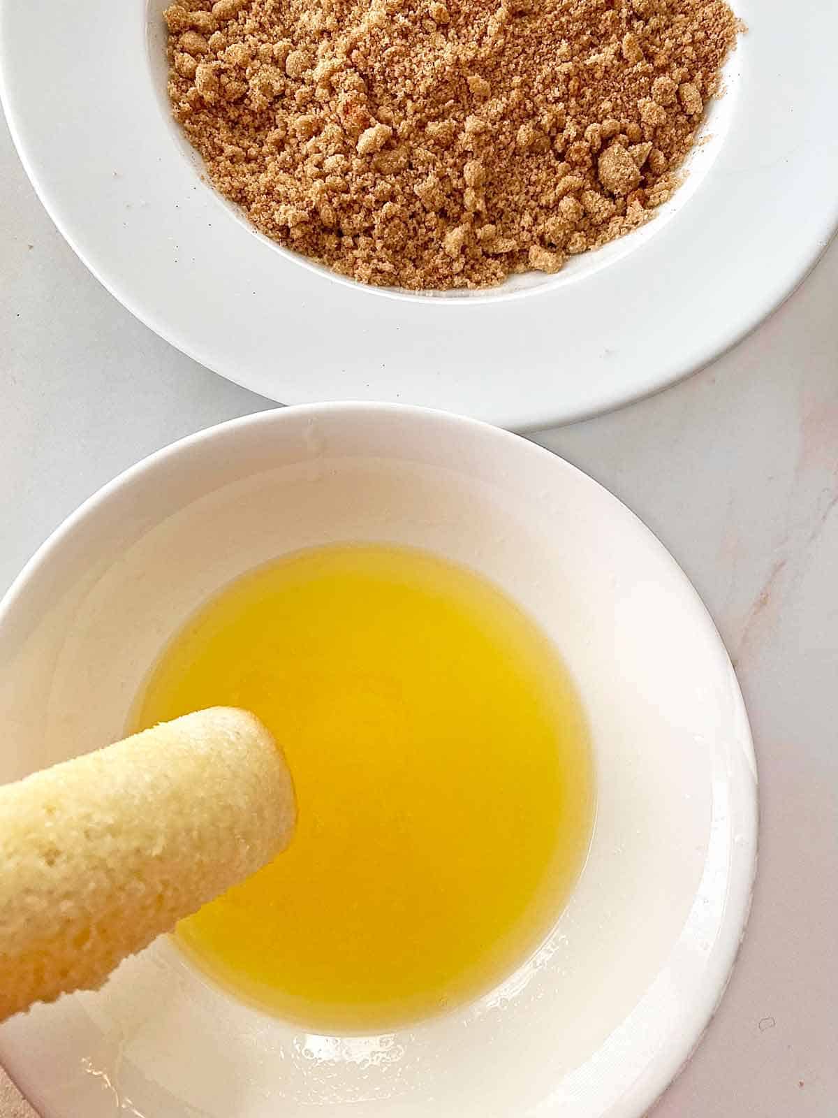 Dipping the cream cheese rollup into melted butter, with a bowl of cinnamon sugar topping nearby.