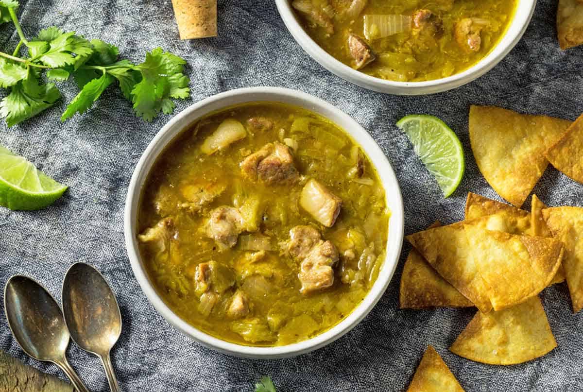 Two bowls of green chile surrounded by tortilla chips and cut limes.