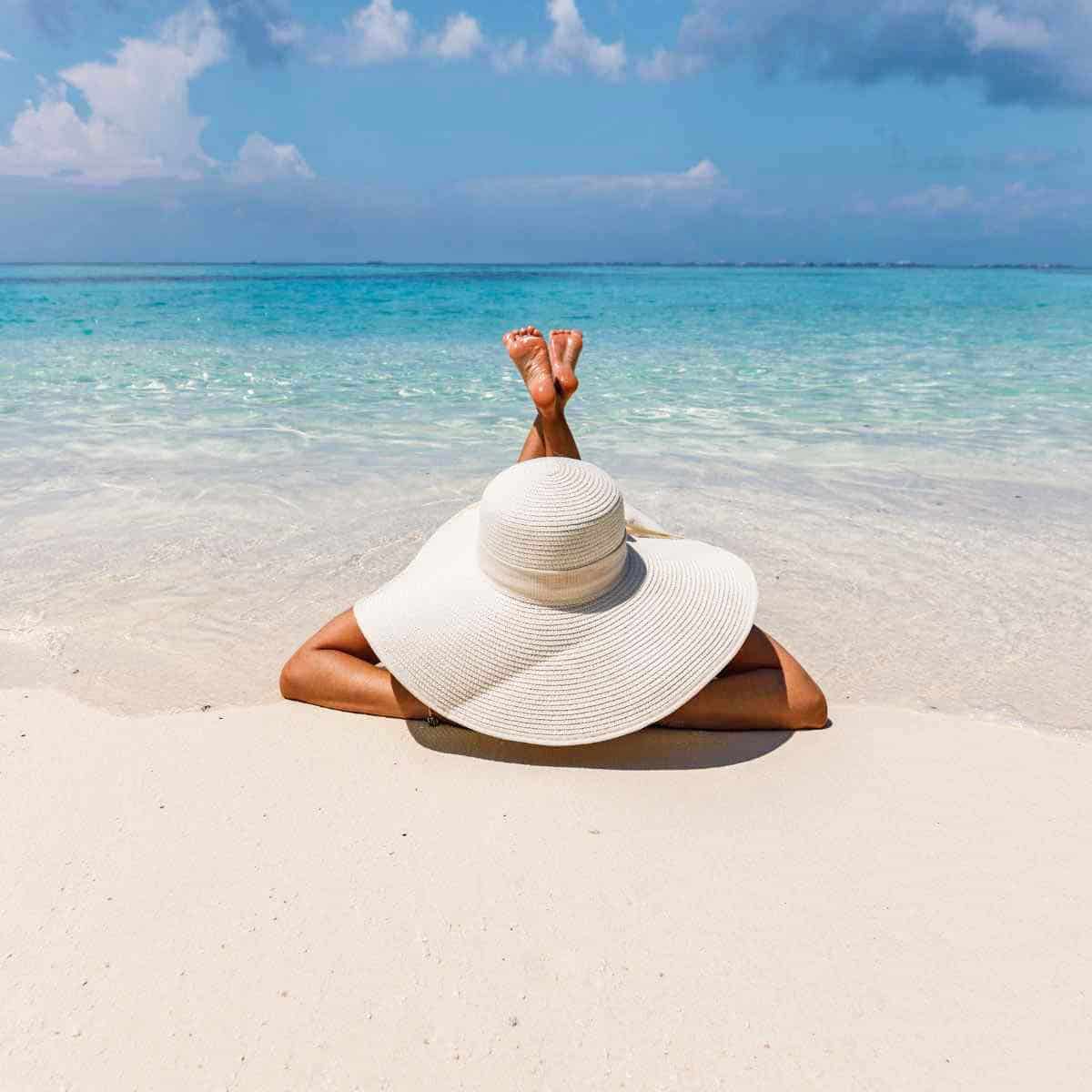 A woman wearing a large sun hat and lying on the beach.