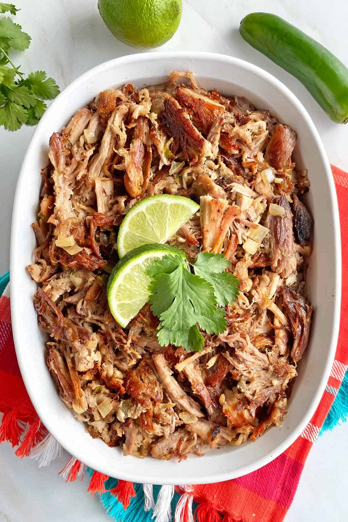 A white serving dish filled with Mexican Carnitas, garnished with limes and cilantro and ready for serving.