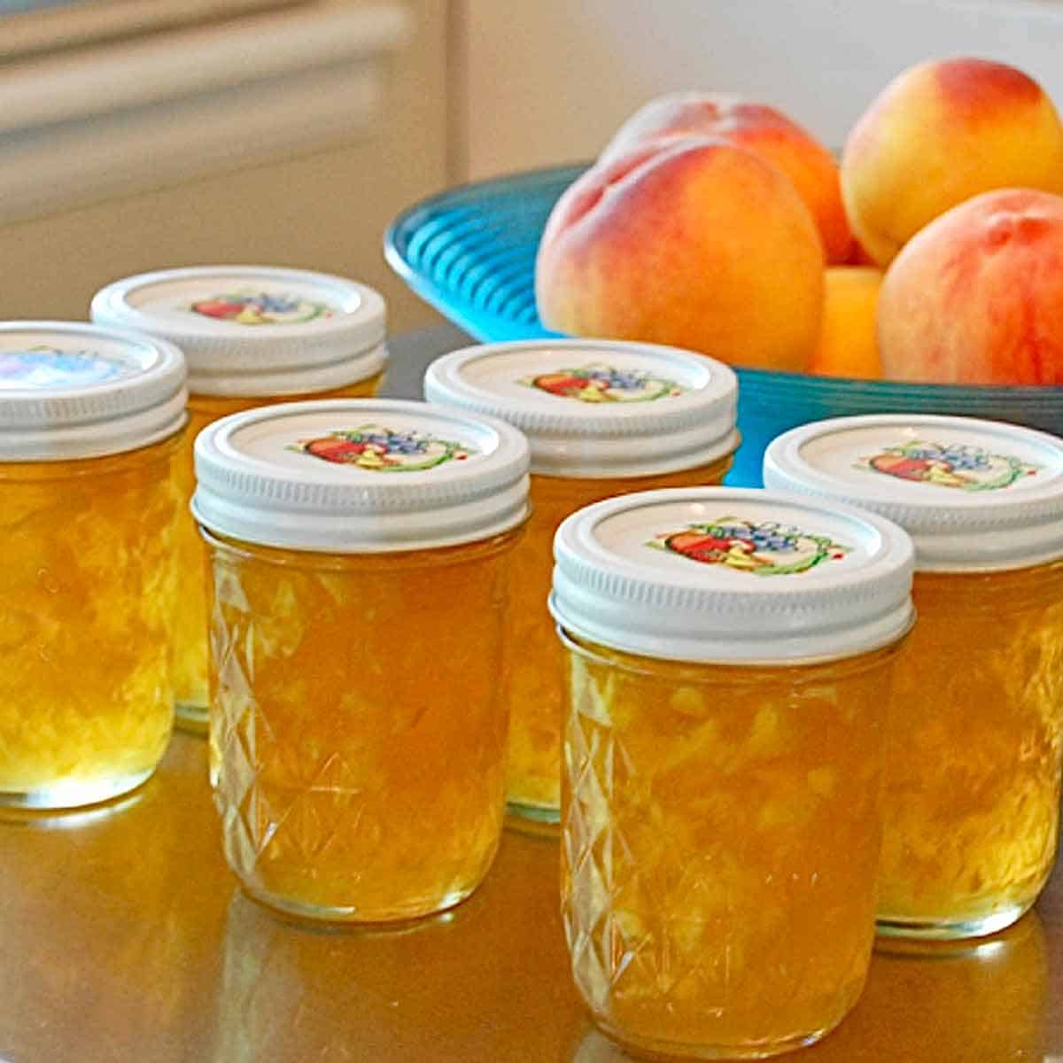 peach jam Eight jars of homemade peach preserves in front of a bowl of fresh peaches.