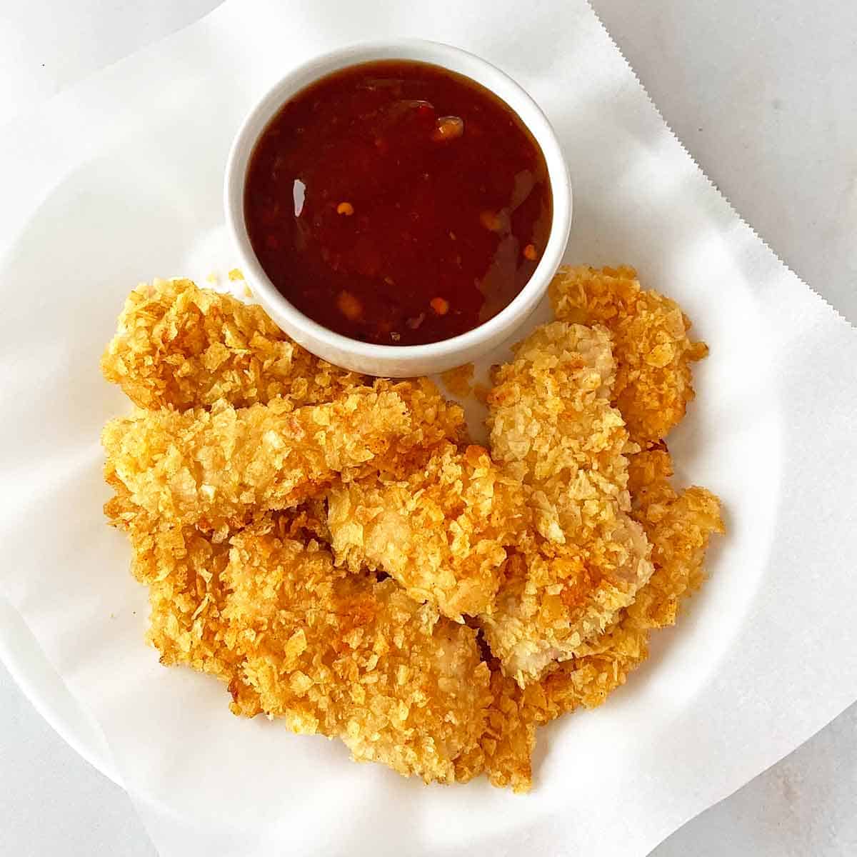 PotatoChipChicken A dozen pieces of golden brown potato chip chicken tenders on a plate with sweet chili sauce for dipping.