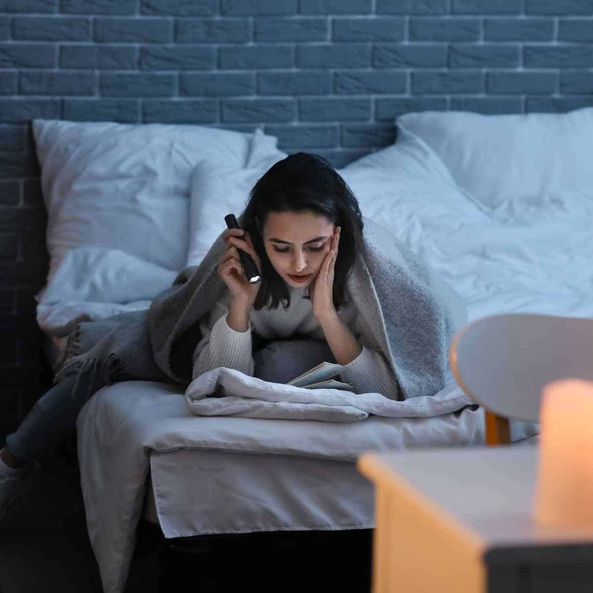 A woman reading a book with a flashlight and candles nearby, in a darkened room.