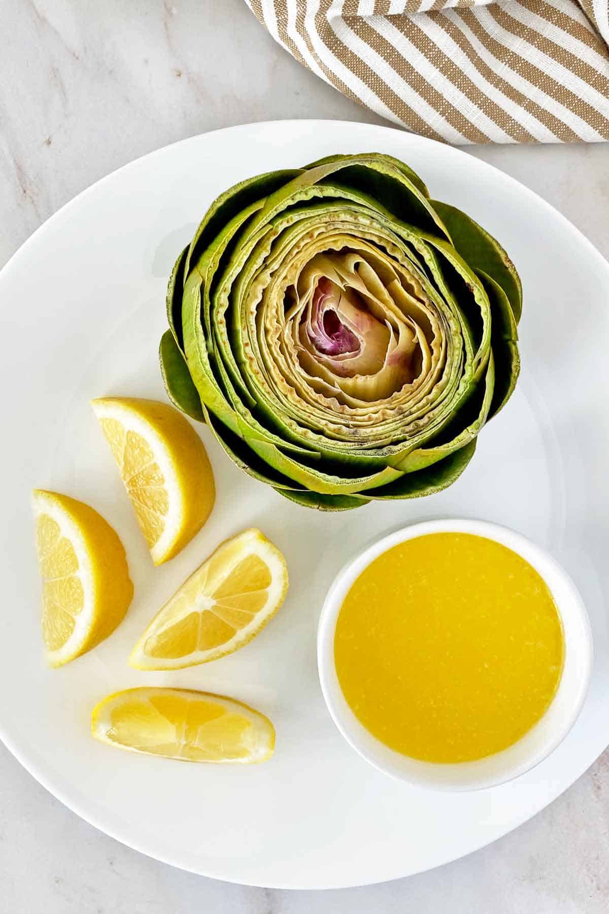 A microwave steamed artichoke on a serving plate with lemon slices and melted butter.
