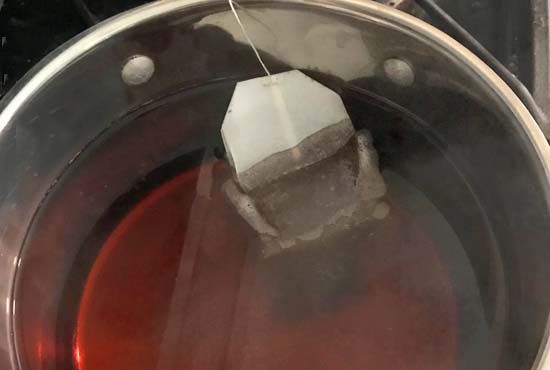 A tea bag being removed from a saucepan filled with hot tea.