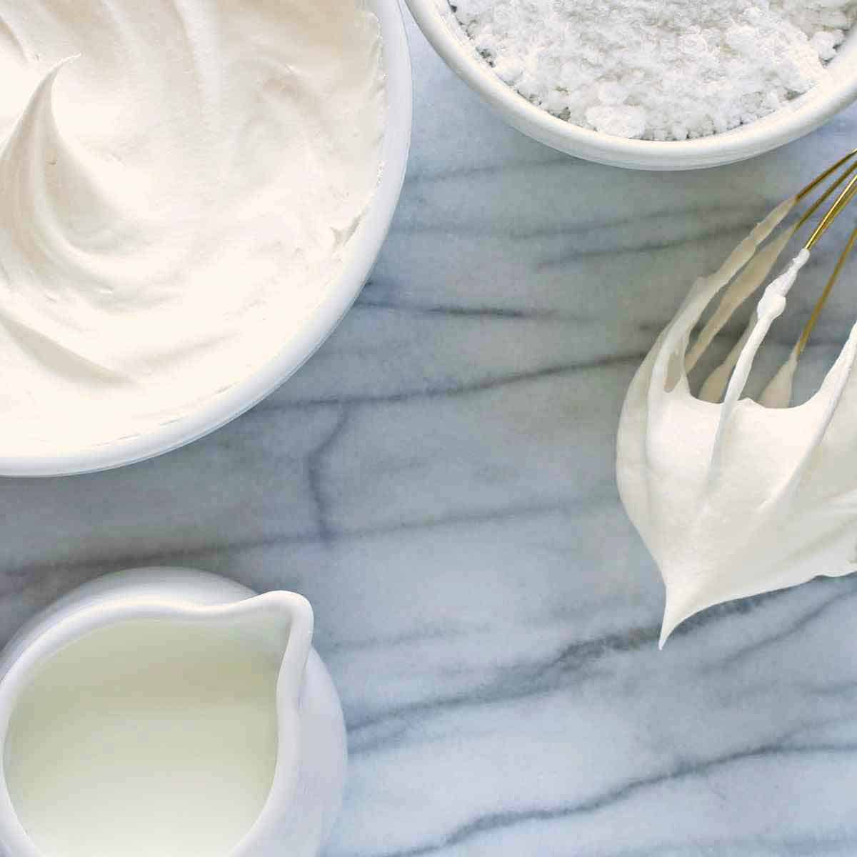 Whipped cream in a bowl with cream, powdered sugar, and a beater nearby.